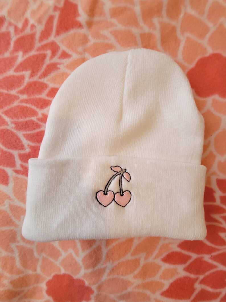 May include: White beanie with a pink cherry embroidered on the front.