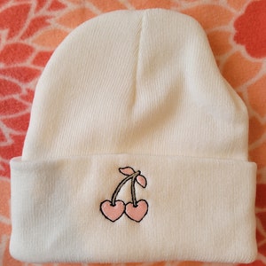 May include: White beanie with a pink cherry embroidered on the front.