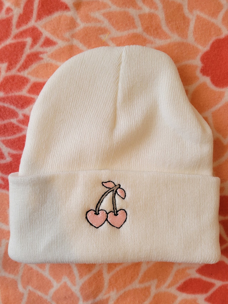 May include: White beanie with a pink cherry embroidered on the front.