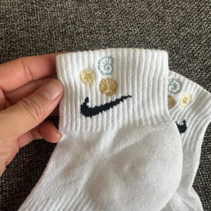 Könnte beinhalten: Weiße Sportsocken mit einem schwarzen Nike Swoosh-Logo und gestickten goldenen und hellblauen Muschelmotiven. Die Socken sind gerippt und scheinen aus einem weichen, atmungsaktiven Material zu sein.