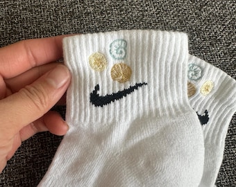 May include: White athletic socks with a black Nike swoosh logo and embroidered gold and light blue seashell designs. The socks are ribbed and appear to be made of a soft, breathable material.