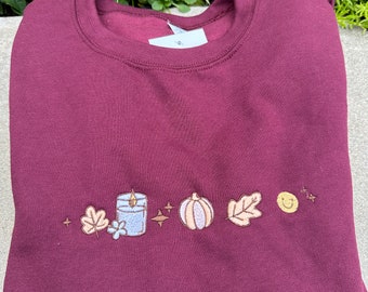 May include: Burgundy sweatshirt with embroidered autumn-themed design. The design features a candle, pumpkin, leaf, and smiley face.