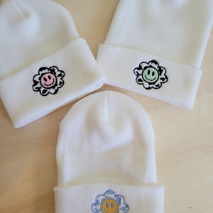 Puede incluir: Tres gorros de punto blanco con dise&ntilde;os de flores bordadas. Cada gorro presenta una cara sonriente de diferente color en el centro de la flor. Los colores de las flores incluyen rosa, verde y azul. Los gorros est&aacute;n doblados en el borde.