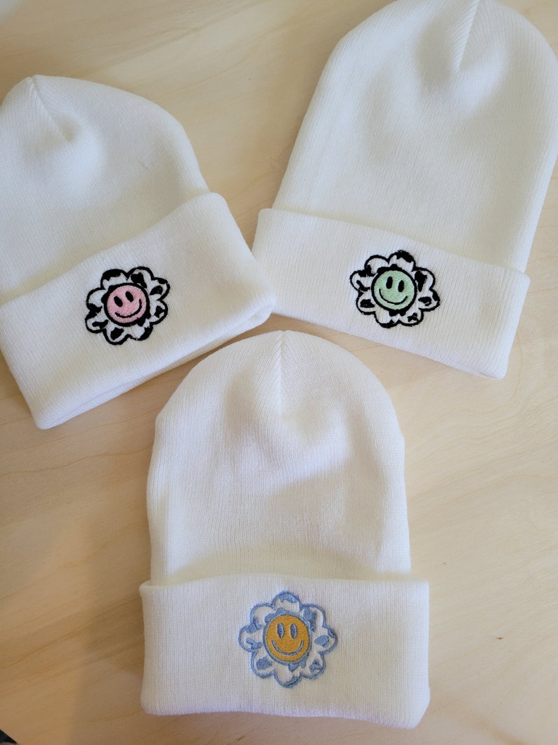 Puede incluir: Tres gorros de punto blanco con dise&ntilde;os de flores bordadas. Cada gorro presenta una cara sonriente de diferente color en el centro de la flor. Los colores de las flores incluyen rosa, verde y azul. Los gorros est&aacute;n doblados en el borde.