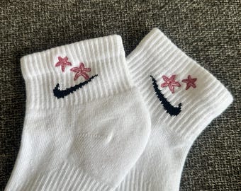 May include: A pair of white ankle socks with a ribbed texture. Each sock features a black swoosh logo and two pink flower designs embroidered above the logo. The socks are made of a soft, comfortable material.
