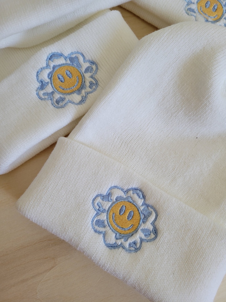Puede incluir: Conjunto de gorro y diadema de color crema. Cada art&iacute;culo presenta un dise&ntilde;o bordado de una carita sonriente amarilla dentro de una flor azul. El gorro tiene un dobladillo doblado. Los art&iacute;culos est&aacute;n hechos de un material suave y texturizado.