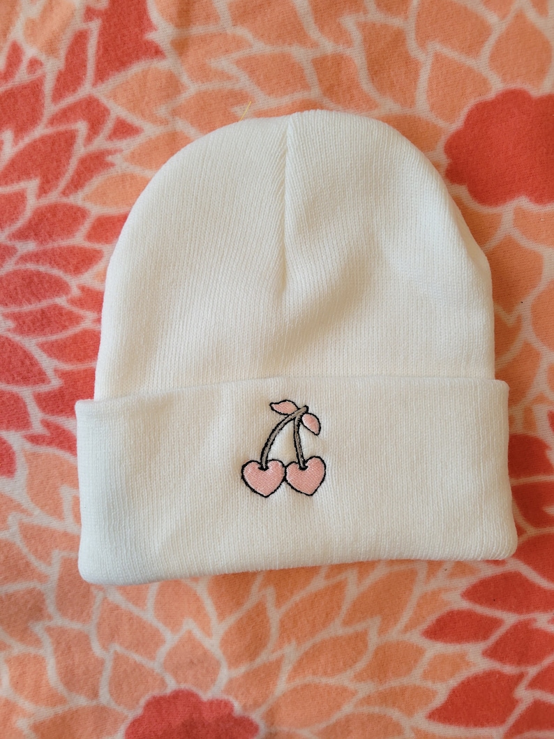 May include: White knitted beanie with a pink cherry embroidered on the front.
