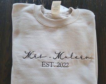 Puede incluir: Una sudadera de color beige claro con el texto "Mrs. Mulero EST. 2022" bordado en negro.