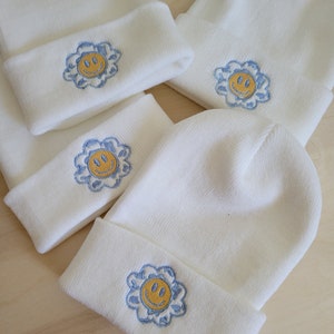 Puede incluir: Varios gorros de punto blancos con dobladillo. Cada gorro presenta un dise&ntilde;o bordado de una cara sonriente amarilla dentro de una flor azul. Los gorros est&aacute;n colocados sobre una superficie de madera clara.