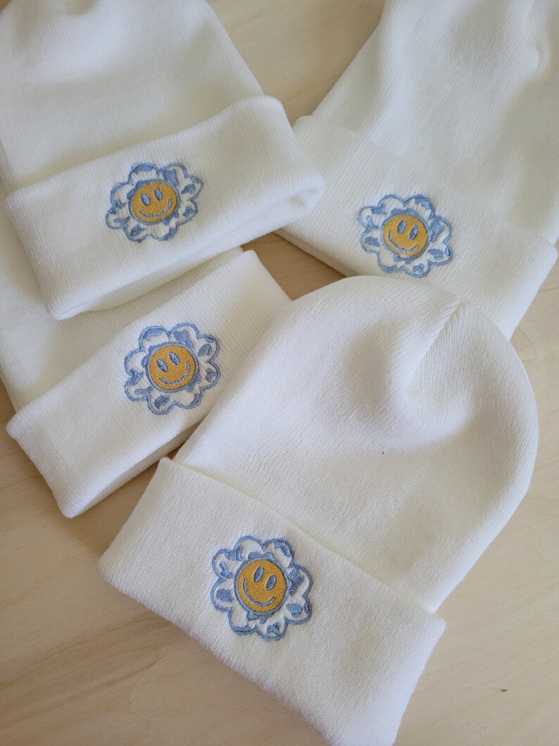 Puede incluir: Varios gorros de punto blancos con dobladillo. Cada gorro presenta un dise&ntilde;o bordado de una cara sonriente amarilla dentro de una flor azul. Los gorros est&aacute;n colocados sobre una superficie de madera clara.