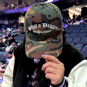 May include: A camouflage baseball cap featuring the white embroidered text What a Dagger above a white embroidered dagger graphic. The cap has a structured crown, a curved brim, and a button at the top center. The fabric displays a green, brown, and black pattern.