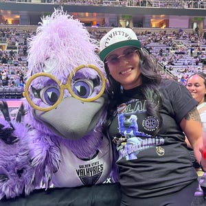 May include: A person poses with the Golden State Valkyries mascot, a purple bird with large glasses. The person wears a black t-shirt with a basketball graphic and a white cap. The background shows a basketball arena.