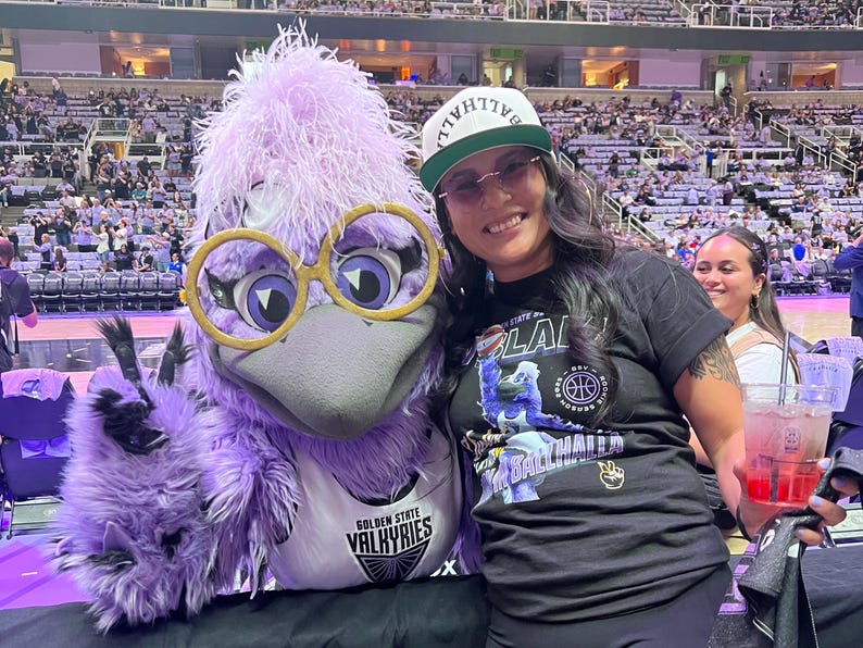 May include: A person poses with the Golden State Valkyries mascot, a purple bird with large glasses. The person wears a black t-shirt with a basketball graphic and a white cap. The background shows a basketball arena.