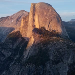 May include: A scenic landscape photograph of a large, imposing rock formation, bathed in the warm glow of the setting sun. The image captures the rugged texture of the rock face and the surrounding mountainous terrain, with a clear blue sky above.