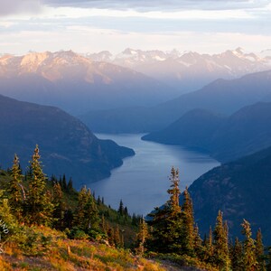 May include: A scenic landscape photograph of a mountain range and a lake. The image features a valley with a lake winding through it, surrounded by mountains. The sky is a mix of blue and white, and the foreground has green trees.