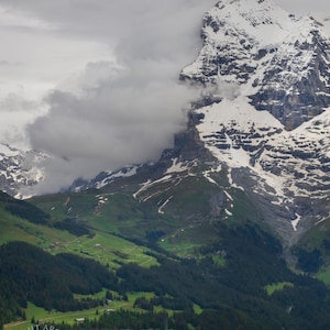 Puede incluir: Un paisaje montañoso con una cima nevada parcialmente oculta por las nubes. Valles verdes exuberantes y densos bosques de hoja perenne llenan el primer plano. La imagen captura una escena natural al aire libre.