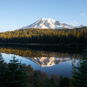 May include: A scenic landscape featuring a snow-capped mountain reflected in a calm lake. The foreground includes evergreen trees and the background is a clear blue sky. The image captures a serene natural environment.