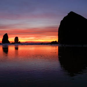 Peut inclure: Un coucher de soleil spectaculaire sur une plage, avec de grandes formations rocheuses en silhouette. Le ciel est peint de couleurs orange, rouge et violet, reflétées dans l'eau, créant une ambiance paisible.