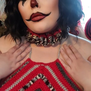 May include: A person wearing a red and gray crocheted halter top with a black and white crocheted collar. The person has black hair and is wearing clown makeup.