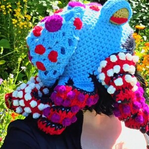 May include: A whimsical, handmade crochet hat in the shape of a sea creature. The hat features a light blue body with red and purple accents, and white and red tentacles. The hat is a unique and colorful accessory.