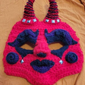 May include: A handmade, red crocheted mask with black and purple accents. The mask features two horns with black stripes, eye shapes, a nose, and a mouth. Silver-colored studs are placed on the horns and cheeks.