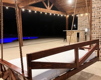 Op de afbeelding: Een houten hangbed met een witte matras hangt aan een overdekt terras met een bakstenen muur en een zwembad op de achtergrond.