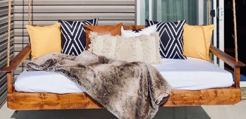 Op de afbeelding: Een houten schommelbed met een wit matras en verschillende kussens. De kussens zijn in tinten bruin, geel en zwart met geometrische patronen. Een nep bontdeken is over het bed gelegd.