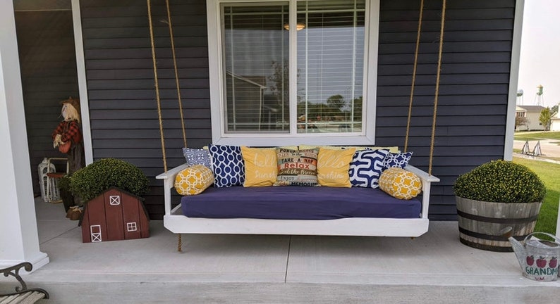 Op de afbeelding: Een witte veranda-schommel met blauwe kussens en gele kussens. De schommel hangt aan een veranda met donkergrijze bekleding. De tekst op een van de kussens is "Take a Nap & Relax".