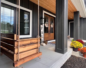 Op de afbeelding: Een houten veranda-schommel hangt aan het plafond van een veranda met een zwart-witte buitenkant. De veranda heeft een houten vloer en een wit plafond. Er zijn herfstdecoraties op de deur, waaronder een krans en een bord met de tekst "FALL".