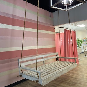 Op de afbeelding: Een witte houten schommelbank hangt aan het plafond met bruine touwen. De schommel bevindt zich voor een roze en witte gestreepte muur. De schommel bevindt zich in een kamer met een roze gordijn op de achtergrond.