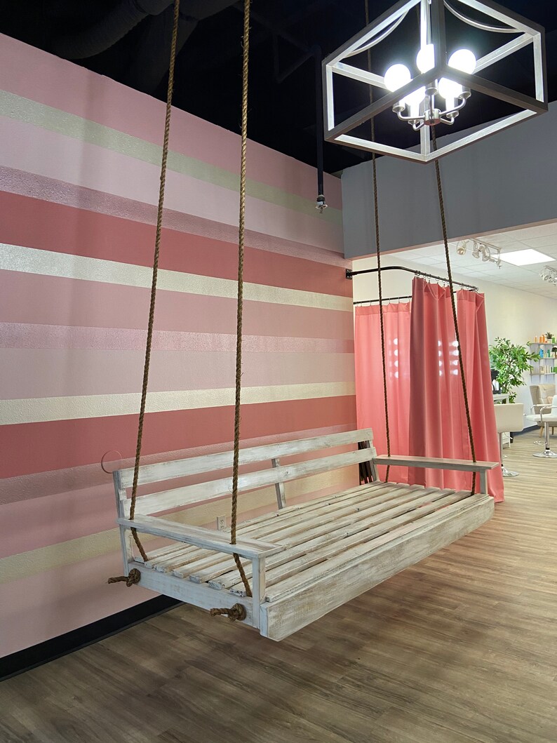 Op de afbeelding: Een witte houten schommelbank hangt aan het plafond met bruine touwen. De schommel bevindt zich voor een roze en witte gestreepte muur. De schommel bevindt zich in een kamer met een roze gordijn op de achtergrond.