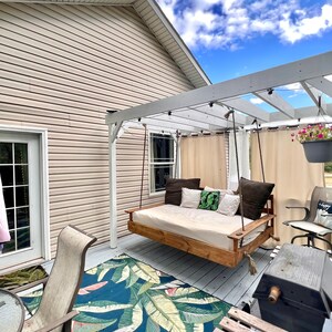 Op de afbeelding: Een wit houten hangbed hangt aan een pergola op een houten dek. Het hangbed heeft een witte matras en bruine kussens. Een blauw en groen tropisch bladmotief tapijt ligt op het dek.