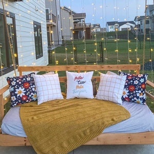 Op de afbeelding: Een houten schommelbed met een wit-blauwe gestreepte matras en een gele deken. Het schommelbed is versierd met kussens, waaronder een met de tekst "Hello Ciao Hola Bonjour". Het schommelbed hangt aan een touw en is omgeven door lichtslingers.