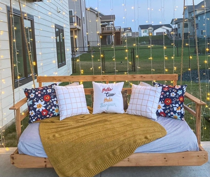 Op de afbeelding: Een houten schommelbed met een wit-blauwe gestreepte matras en een gele deken. Het schommelbed is versierd met kussens, waaronder een met de tekst "Hello Ciao Hola Bonjour". Het schommelbed hangt aan een touw en is omgeven door lichtslingers.