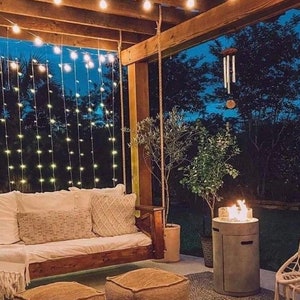 Op de afbeelding: Een knusse buitenpatio met een houten pergola, lichtslingers en een hangende schommel. Er zijn twee bruine poefs, een vuurplaats en een potplant.