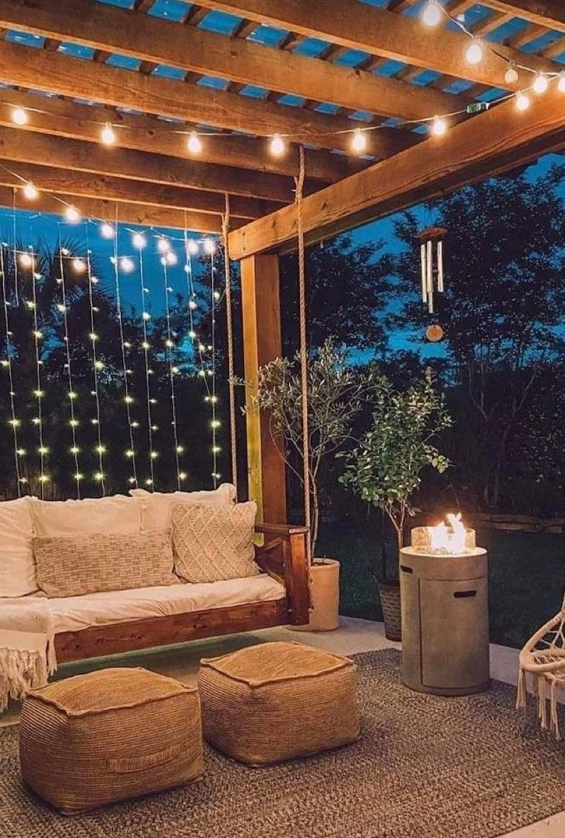 Op de afbeelding: Een knusse buitenpatio met een houten pergola, lichtslingers en een hangende schommel. Er zijn twee bruine poefs, een vuurplaats en een potplant.