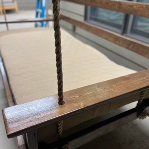 Op de afbeelding: Een houten schommelbed met een bruine beitsafwerking en een beige kussen. De schommel wordt opgehangen aan dikke, bruine touwen.