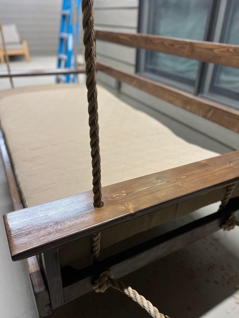 Op de afbeelding: Een houten schommelbed met een bruine beitsafwerking en een beige kussen. De schommel wordt opgehangen aan dikke, bruine touwen.