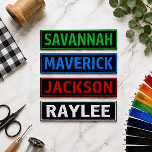 Könnte beinhalten: Vier rechteckige, bestickte Namensaufnäher mit schwarzem Hintergrund und farbigen Rändern. Die Aufnäher zeigen die Namen SAVANNAH in Grün, MAVERICK in Blau, JACKSON in Rot und RAYLEE in Weiß, jeweils in einer fetten, serifenlosen Großschrift.