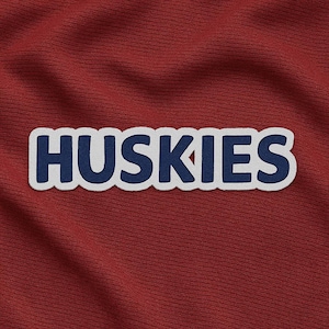 May include: A close-up of a maroon fabric with the word "Huskies" embroidered in navy blue with a white border. The fabric has a textured appearance.