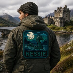May include: An olive green hooded jacket featuring a large square patch on the back. The patch displays a dark green sea creature in water under a full moon, with the text "NESSIE" printed in bold, light-colored capital letters at the bottom.