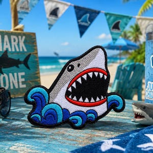 May include: Embroidered shark patch with a gray body, white teeth, and red mouth, set against blue waves. The patch is on a blue wooden surface with sunglasses, a sign that says "Shark Zone", and a shark-themed tumbler.