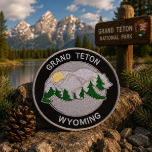May include: A circular embroidered patch featuring a black border with the white text GRAND TETON at the top and WYOMING at the bottom. The center displays a light gray bear silhouette containing a yellow sun and green pine tree graphics.