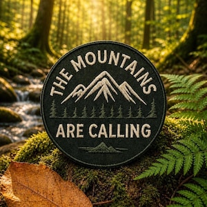 May include: A circular dark green embroidered patch featuring a white mountain range graphic, a line of small pine trees, and the text "THE MOUNTAINS ARE CALLING" in a bold, sans-serif font, with a stitched border edge.