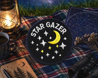 May include: A black patch with the words "STAR GAZER" in white, along with white stars and a yellow crescent moon. The patch is on a plaid blanket, with a lantern, coffee mug, notebook, and binoculars in the background.