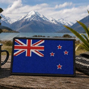 May include: A rectangular blue patch displaying the New Zealand flag, with a red Union Jack and four red stars. Beside it is a mug with the text "Adventure Awaits" and a mountain design. A signpost indicates directions to Roy's Peak and other locations.