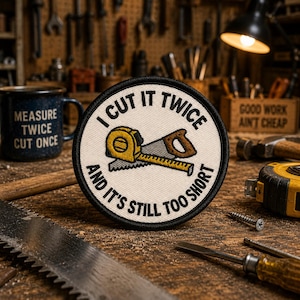May include: A circular patch with the text "I CUT IT TWICE AND IT'S STILL TOO SHORT" surrounds a yellow tape measure and a brown-handled saw. The patch is on a wooden surface with tools and a mug that says "MEASURE TWICE CUT ONCE".