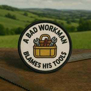 May include: Round patch with the text "A BAD WORKMAN BLAMES HIS TOOLS" in a circular design. The patch features a yellow toolbox with wrenches and a screwdriver. The patch has a black border and is set against a blurred green background.