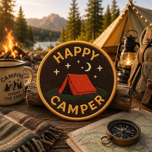 May include: A circular embroidered patch featuring a dark brown background with a red tent on green ground. The text HAPPY CAMPER is stitched in yellow, accompanied by a yellow crescent moon and small stars above the tent design.