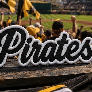 May include: A black and white embroidered patch featuring the word Pirates in a cursive script font. The patch has a thick white border and a textured black interior, designed to be applied to fabric surfaces as a decorative sports team emblem.